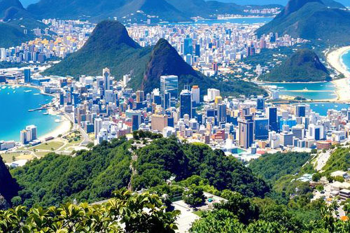 Photo of the beautiful city of Rio de janeiro, beautiful lush green mountains with buildings between them, and icy blue waters.